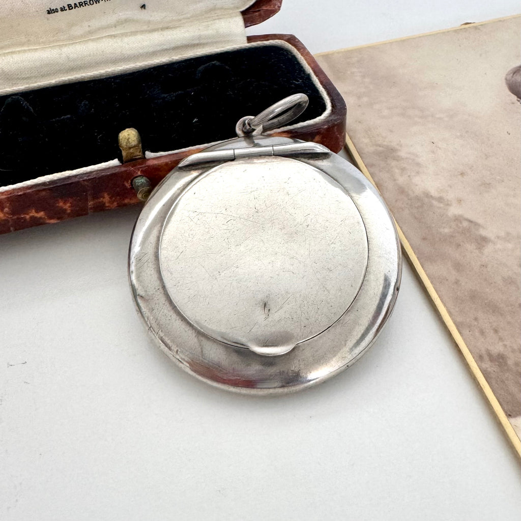 A round antique silver compact mirror with a hinged lid and top loop, shown open on a white surface next to its vintage brown leather case.
