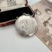 An antique Adie & Lovekin round silver toned compact pendant with a small loop at the top, shown resting on a white surface beside an open vintage presentation box and an old sepia-toned portrait photograph.