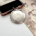 An antique silver oval compact mirror with a hinged lid, displayed next to its open brown leatherette case and an old sepia-toned photograph.