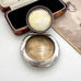 An antique Adie & Lovekin open round silver compact or powder case with a hinged lid, shown on a light surface beside its dark velvet-lined wooden box and an old printed page. The inside of the lid has a pale cream-colored insert or cover.