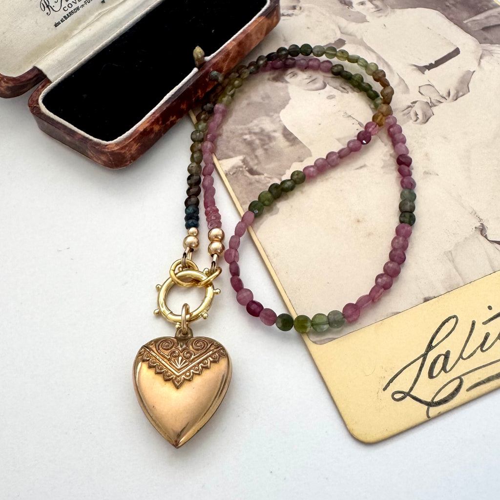A beaded tourmaline necklace with an antique Etruscan revival 9ct gold lined heart pendant, displayed on a vintage-style card next to an open jewelry box. The beads transition from dark green to pinkish-purple, and the pendant is gold-toned with engraved details. 