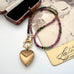 A beaded tourmaline necklace with an antique Etruscan revival 9ct gold lined heart pendant, displayed on a vintage-style card next to an open jewelry box. The beads transition from dark green to pinkish-purple, and the pendant is gold-toned with engraved details. 