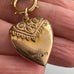 A close-up of an antique gold heart-shaped  pendant held in a hand. The pendant features an engraved top section with scrollwork and a decorative border, and the word 9ct lined is stamped on the lower right side.