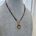 A beaded necklace with a heart-shaped pendant displayed on a gray fabric mannequin bust. The necklace features small faceted beads in pink, green, and black, connected by gold-toned metal links, with a gold heart pendant hanging from a ring clasp.
