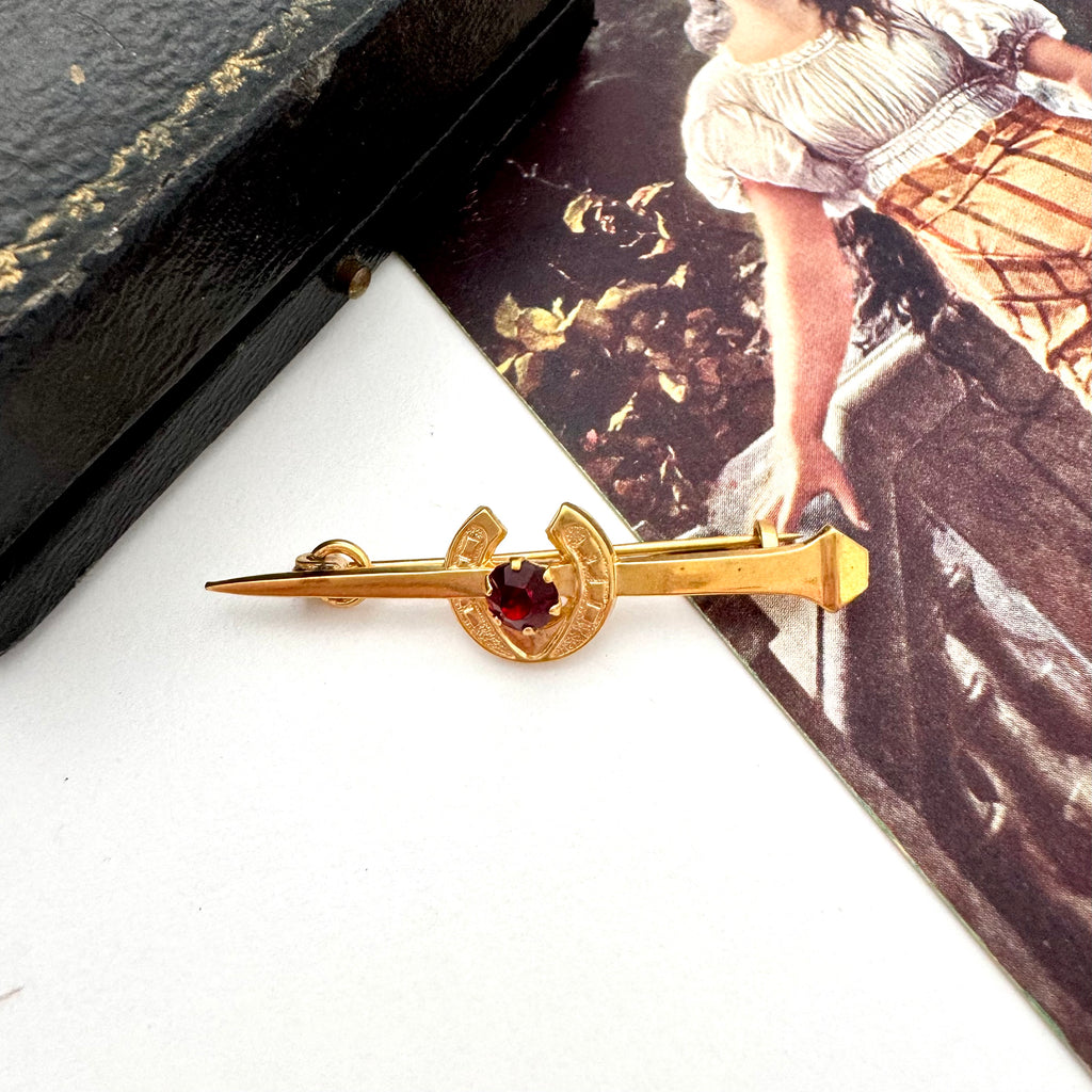 Vintage 9ct gold lined horseshoe and nail brooch with a red gemstone on a white surface with a blurred background