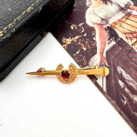 Vintage 9ct gold lined horseshoe and nail brooch with a red gemstone on a white surface with a blurred background