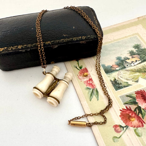 Antique Edwardian stanhope Lourdes binoculars necklace on a chain with a black case and floral illustration in the background.