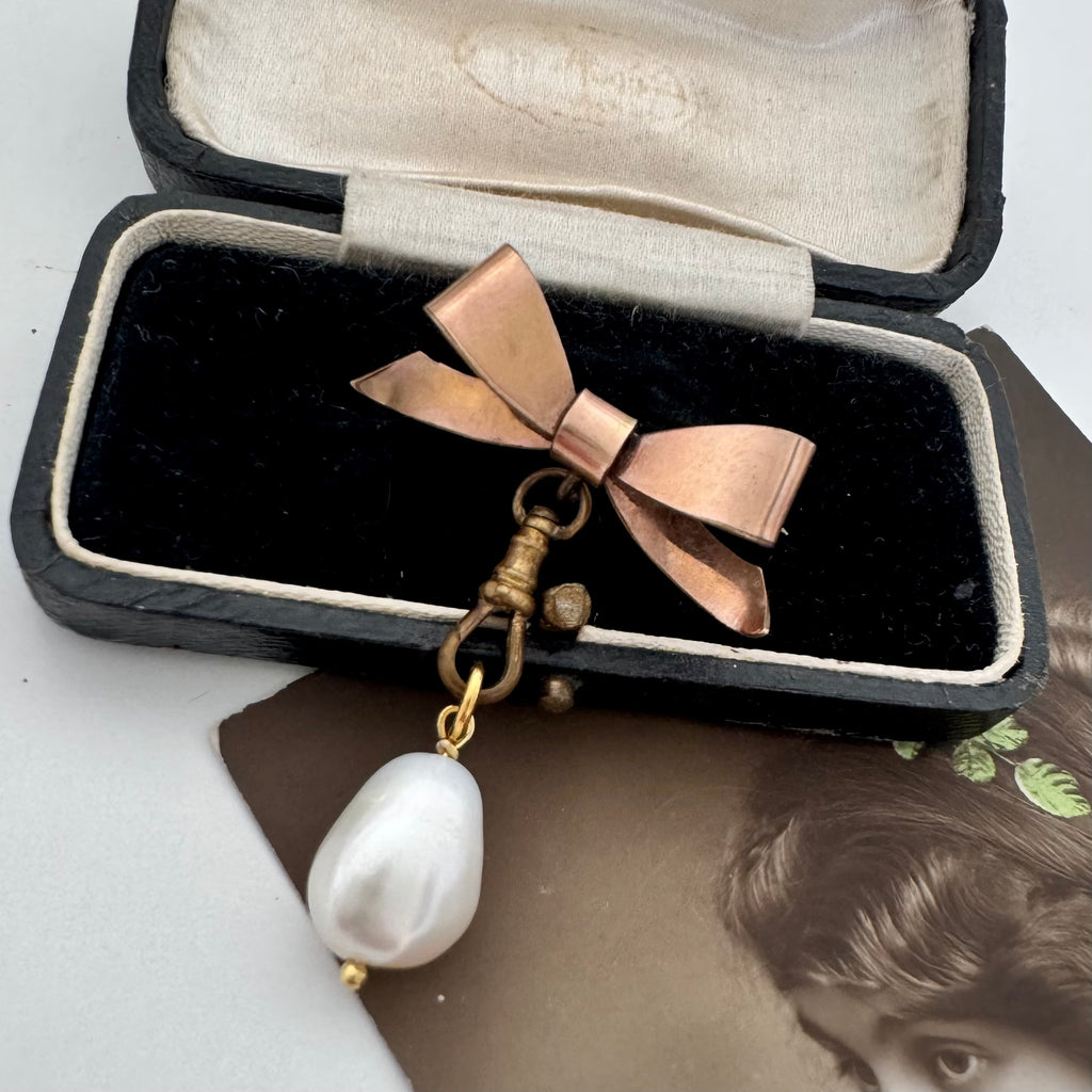 An Edwardian rolled rose gold bow-shaped brooch with a baroque pearl drop and brass clasp, displayed inside an open vintage black jewelry box.