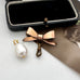 An Edwardian rolled rose gold bow-shaped brooch with a dangling clasp and a white baroque pearl pendant on a gold loop, displayed next to an open black jewelry box on a white surface.