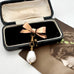 An Edwardian rolled gold bow brooch, gold-toned clasp, and a dangling white baroque pearl, displayed inside an open black presentation box with a cream velvet lining.