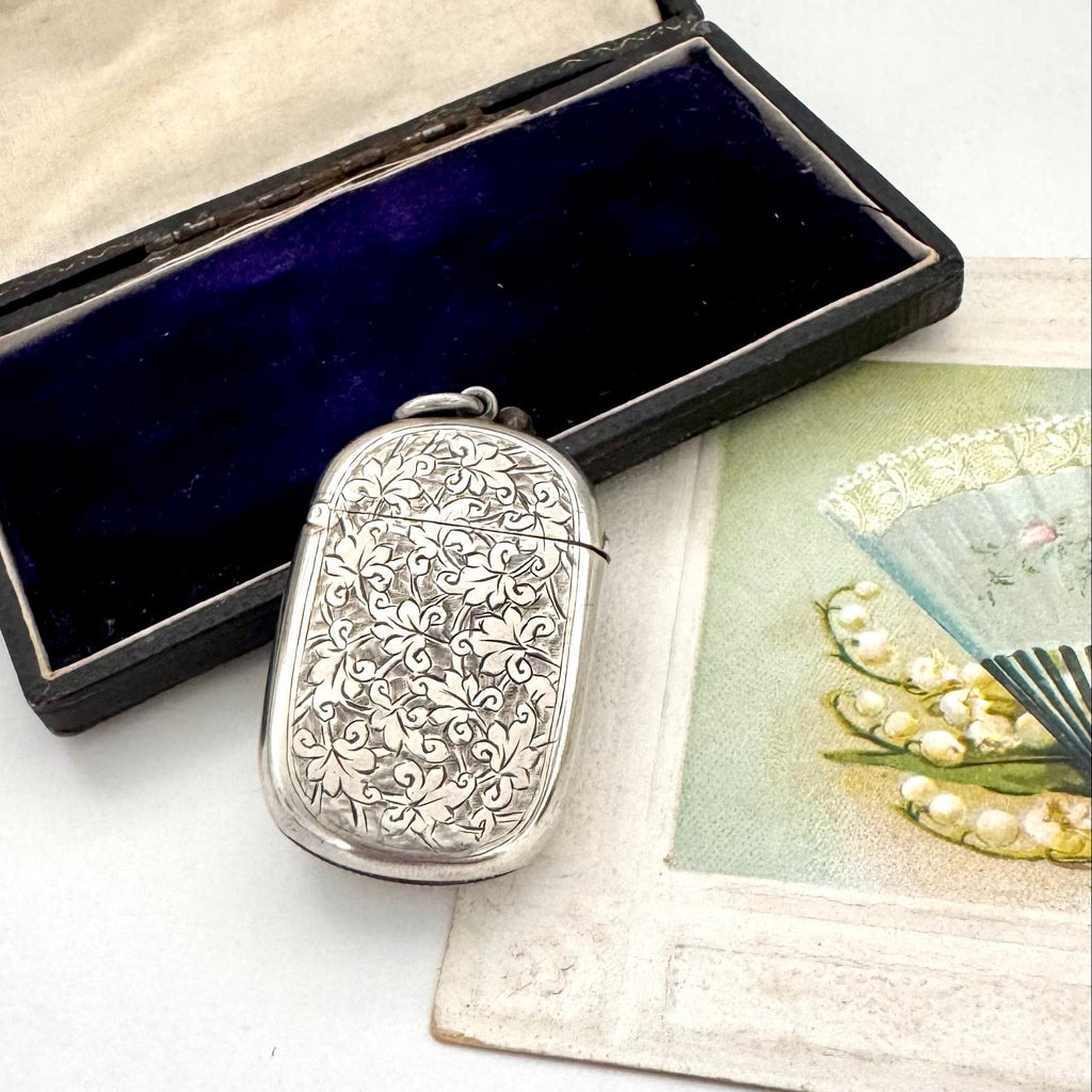 Antique sterling silver 1887 vesta case with intricate patterns on a white surface next to an open jewelry box and vintage card.