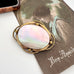 An antique oval brooch featuring a iridescent mother-of-pearl center with a gold-toned metal border, displayed on a vintage-style print over a white surface.
