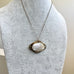 A delicate chain necklace with an antique oval pendant featuring a white mother-of-pearl center and a gold-toned metal border, displayed on a light gray fabric jewelry bust.