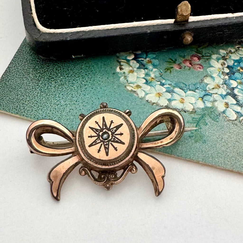 Antique Victorian star burst and bow-shaped brooch  displayed on a white surface with a floral card and a black box in the background.