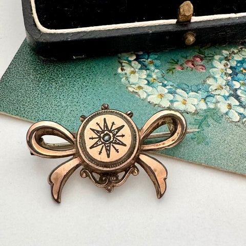Antique Victorian star burst and bow-shaped brooch  displayed on a white surface with a floral card and a black box in the background.