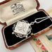 A silver locket pendant with floral engraving, presented in an open vintage wooden box with black velvet lining. The box lid shows printed branding for R.