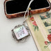 A silver-toned pendant necklace with a square iridescent shell inlay featuring a black ink sketch of a bird, displayed next to an open vintage jewelry box and a floral card.