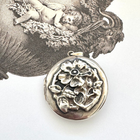 Sterling silver art nouveau repousse floral locket on a white background with an engraved cherub design in the background.