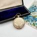 Antique pocket watch style locket with intricate design on a white surface next to an open jewelry box.