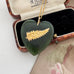 A heart-shaped dark green jade pendant necklace with a gold fern leaf and 'NZ' engraving, displayed inside an open vintage Barrow & Birkham jewelry box.