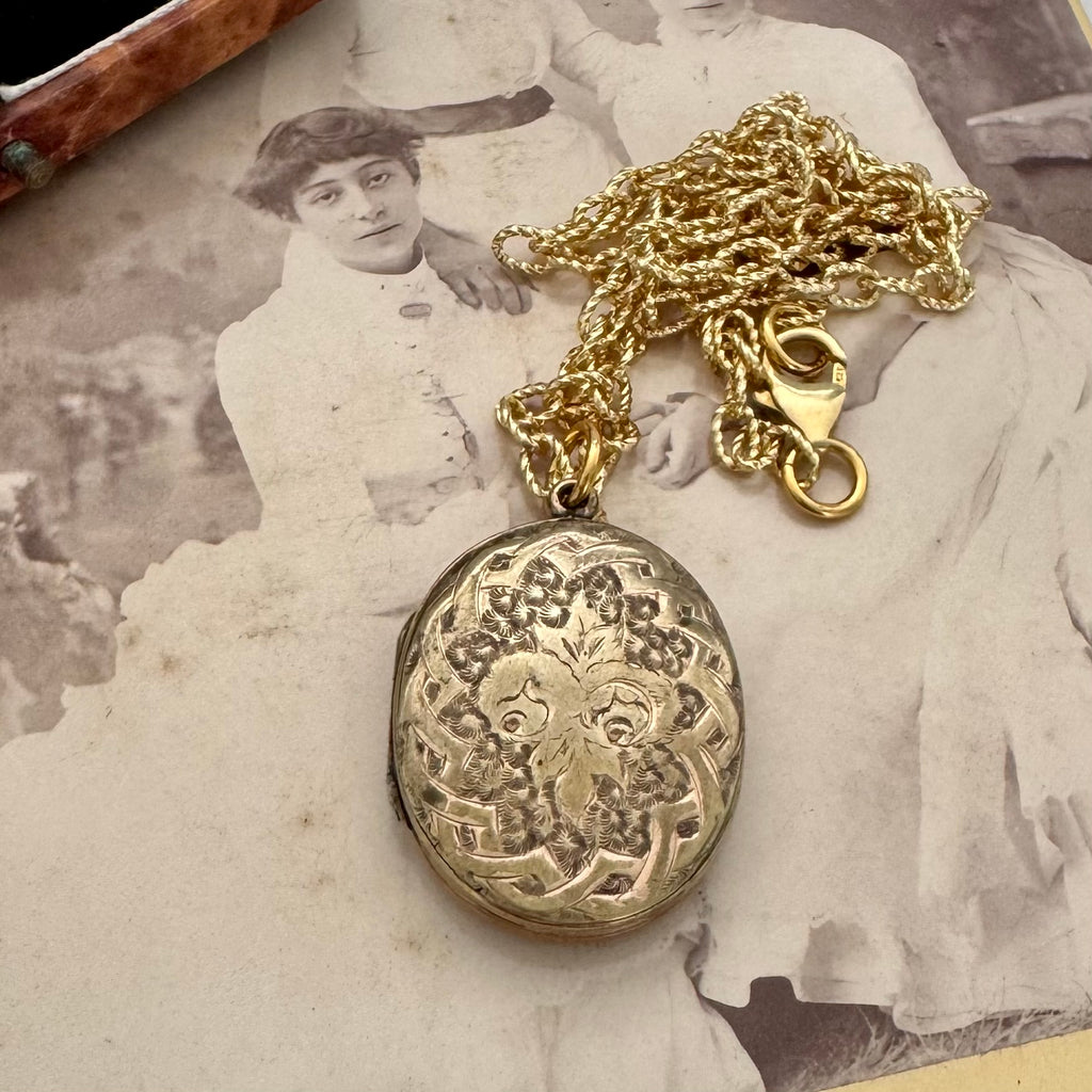 An antique oval locket pendant necklace with an ornate floral engraving, resting on a vintage sepia-toned photograph of a woman. The locket is attached to a gold-tone chain with a lobster clasp.