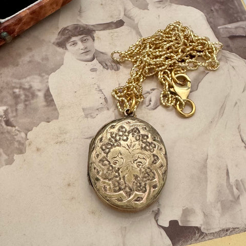An antique oval locket pendant necklace with an ornate floral engraving, resting on a vintage sepia-toned photograph of a woman. The locket is attached to a gold-tone chain with a lobster clasp.