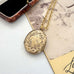 An antique oval gold locket pendant necklace with intricate floral engravings, resting on a vintage-style printed card next to an open jewelry box.