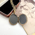 An antique oval locket necklace with dark glass panels and a gold-toned chain, displayed on a vintage photo print next to an open black velvet jewelry box.