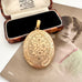 A Victorian oval gold locket pendant with floral engravings, displayed in front of an open vintage presentation box on top of an antique portrait photograph.