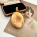 An oval gold pendant locket with a tarnished, aged patina is displayed on top of its original vintage presentation. The locket is resting on an antique sepia-toned portrait print, with its open wooden case featuring a black velvet interior visible behind it.