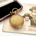 An antique rolled gold sovereign holder with a textured circular cover, displayed next to its open wooden presentation box with black velvet lining, with a vintage portrait photo in the background.