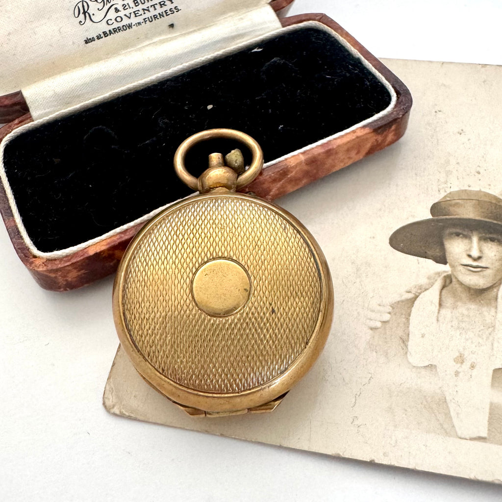 An antique rolled gold sovereign holder with a textured circular cover, displayed next to its open wooden presentation box with black velvet lining, with a vintage portrait photo in the background.