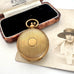 An antique rolled gold sovereign holder with a textured circular cover, displayed next to its open wooden presentation box with black velvet lining, with a vintage portrait photo in the background.