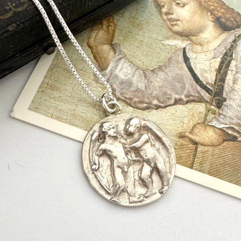 Antique silver pendant necklace on a gray surface with an old book and illustration in the background. The pendant features Cupid and Psyche in relief and has been converted from an antique button.