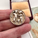 Antique silver button with relief sculpture in a woman's hand and with a wooden box on a white surface.