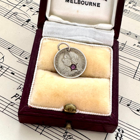 Silver antique threepence coin charm with a purple gemstone in an open jewelry box on sheet music.
