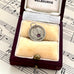 Silver antique threepence coin charm with a purple gemstone in an open jewelry box on sheet music.