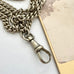 A close-up of a chunky silver plated guard chain necklace with a lobster clasp pendant, resting on a white surface next to a gold card.