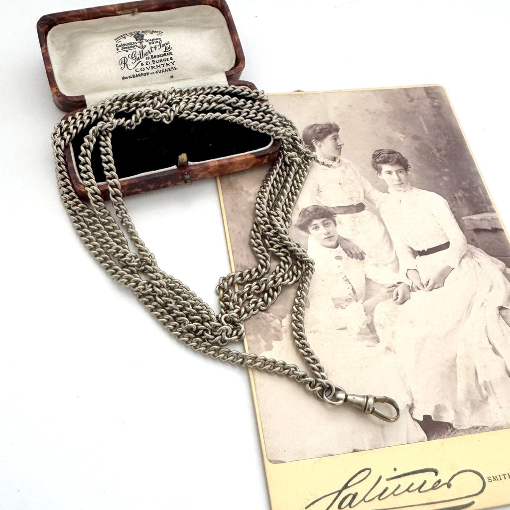 A vintage silver plated guard chain necklace draped over an open brown presentation box, placed on top of an antique black-and-white portrait photograph. 