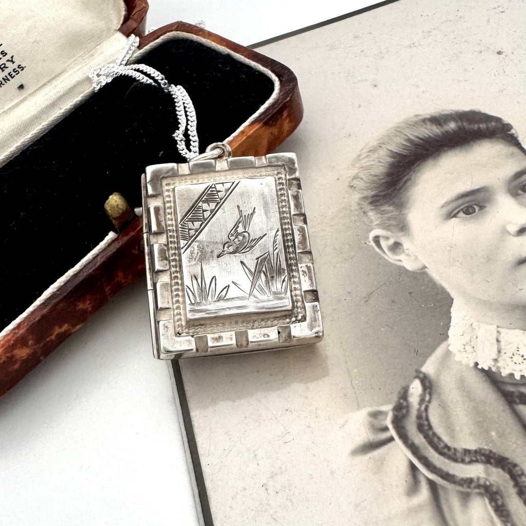 An antique aesthetic era rectangular silver locket with an engraved bird and reeds design, displayed next to its vintage presentation box on top of an antique black-and-white portrait photograph.
