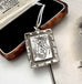 A silver pendant necklace with an engraved monogram for Auspice Mary, resting on its brown velvet-lined presentation box. The box lid shows the brand name 'Reginald & Barbour Coventry'.