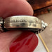 A close-up of a silver-toned ring being held between fingers with red nail polish. The ring has engraved text on its band and decorative side details.