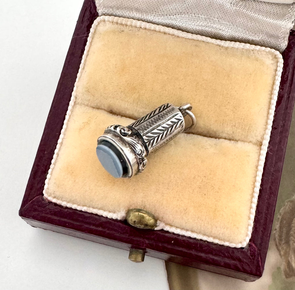 An antique miniature silver wax seal fob  with engraved leaf-like detailing and a round pale blue stone, displayed inside an open burgundy jewelry box with cream velvet lining.