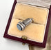An antique miniature silver wax seal fob  with engraved leaf-like detailing and a round pale blue stone, displayed inside an open burgundy jewelry box with cream velvet lining.
