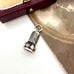An antique miniature wax seal fob with engraved leaf-like detailing, shown on a light surface with a metallic gold-toned sheet and a dark wooden box in the background.