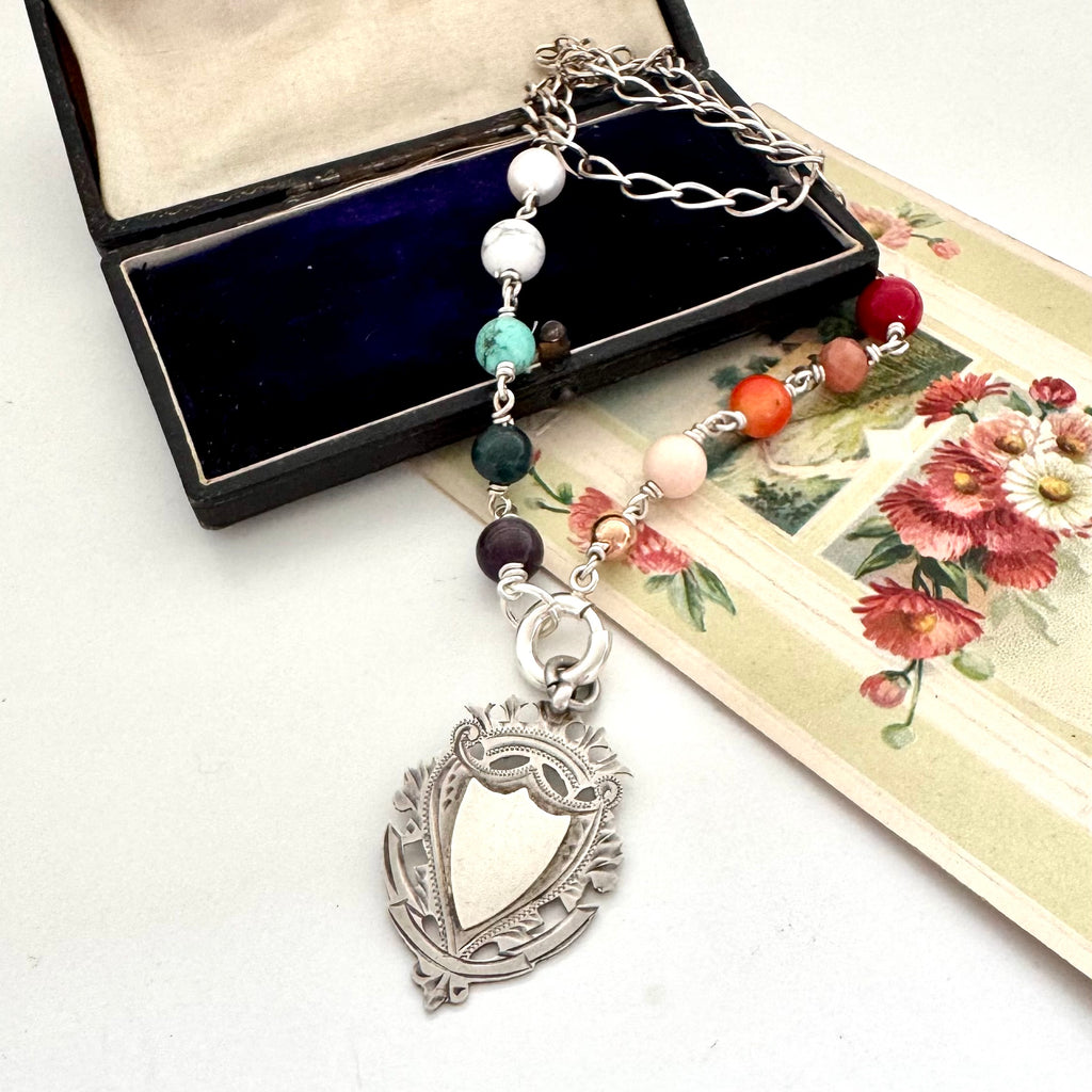 Necklace with multicolored beads and antique silver watch fob on a white surface with an open jewelry box and floral card.