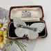 Back of Art Deco era butterfly-shaped brooch with a silver-toned metal pin back, displayed inside an open antique presentation box. The box has a brown wood-grain exterior and a cream-colored interior with printed branding for R.