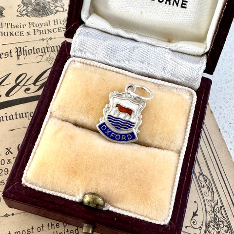 Silver charm with 'Oxford' on a beige pillow in an open jewelry box.