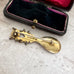 Vintage gold over silver Sami spoon brooch on a textured surface with an open jewelry box in the background.