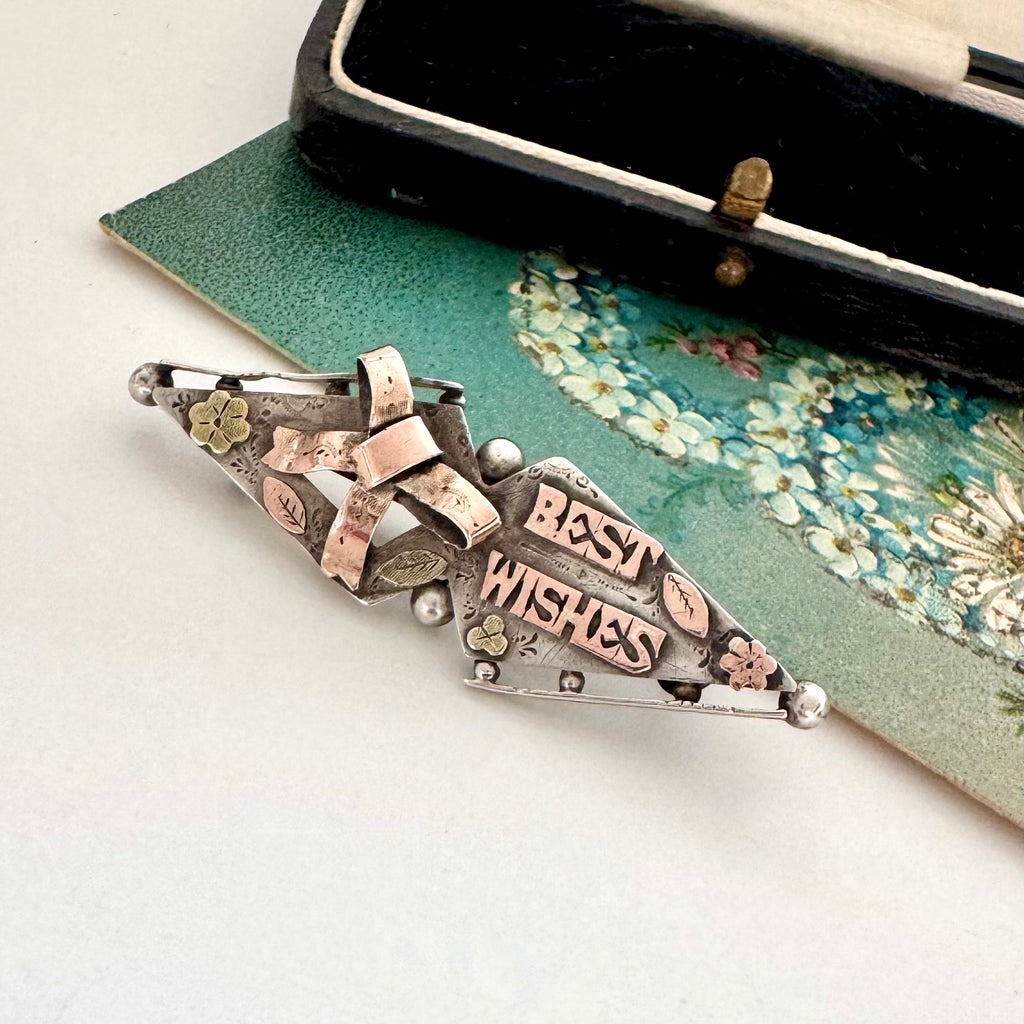 Victorian 1900 antique sterling silver and rose gold sweetheart brooch with the words 'BEST WISHES' in raised lettering, styled on a floral green card next to a vintage black case.